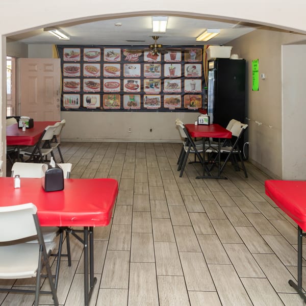 A casual restaurant interior with red tables, white folding chairs, and a large menu board on the back wall.