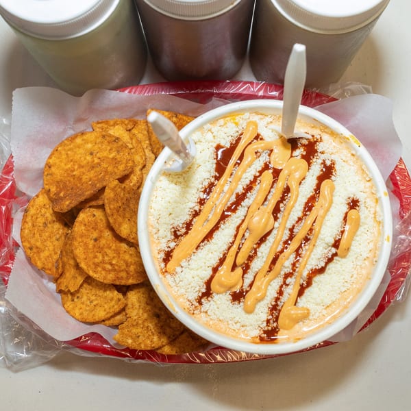 Elote in a white bowl with red and orange sauce, next to a basket of tortilla chips.