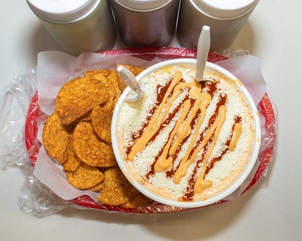 Elote in a white bowl with red and orange sauce, next to a basket of tortilla chips.