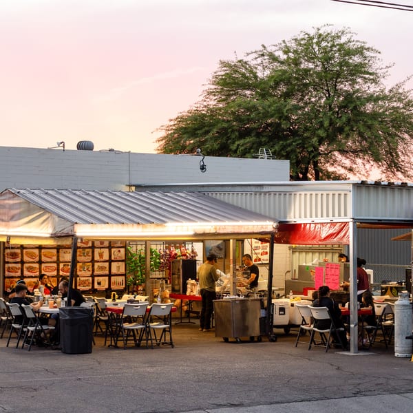 Outdoor restaurant with patrons dining under a lit canopy at dusk, with a large tree and pink sky.