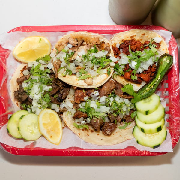 A red basket holds five tacos with various meats, onions, cilantro, lemons, cucumbers, and a grilled jalapeño.