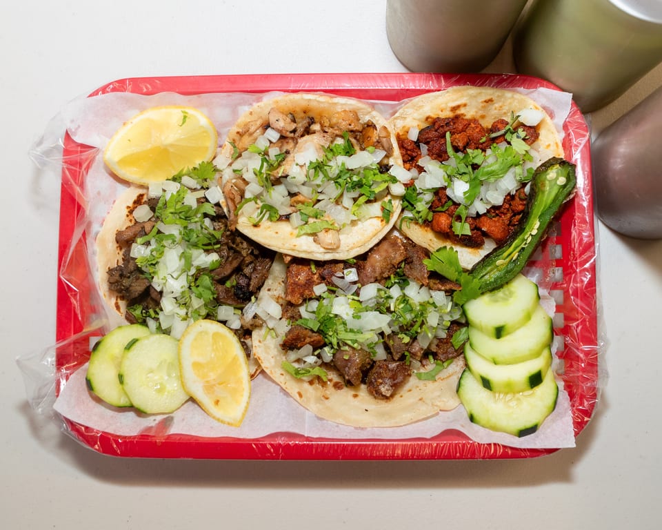 A red basket holds five tacos with various meats, onions, cilantro, lemons, cucumbers, and a grilled jalapeño.