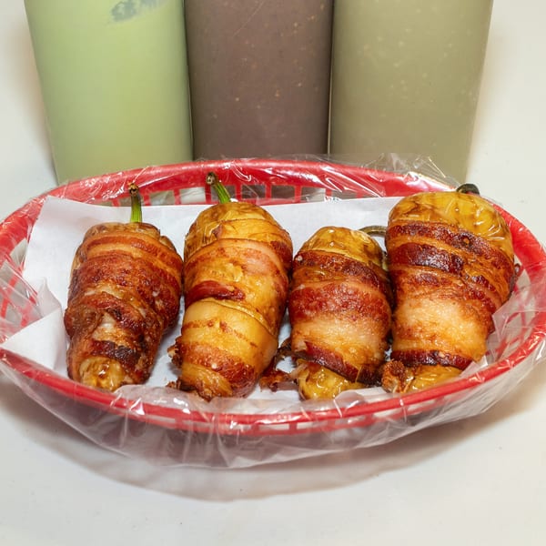 Four bacon-wrapped peppers in a red plastic basket with three drinks in the background.