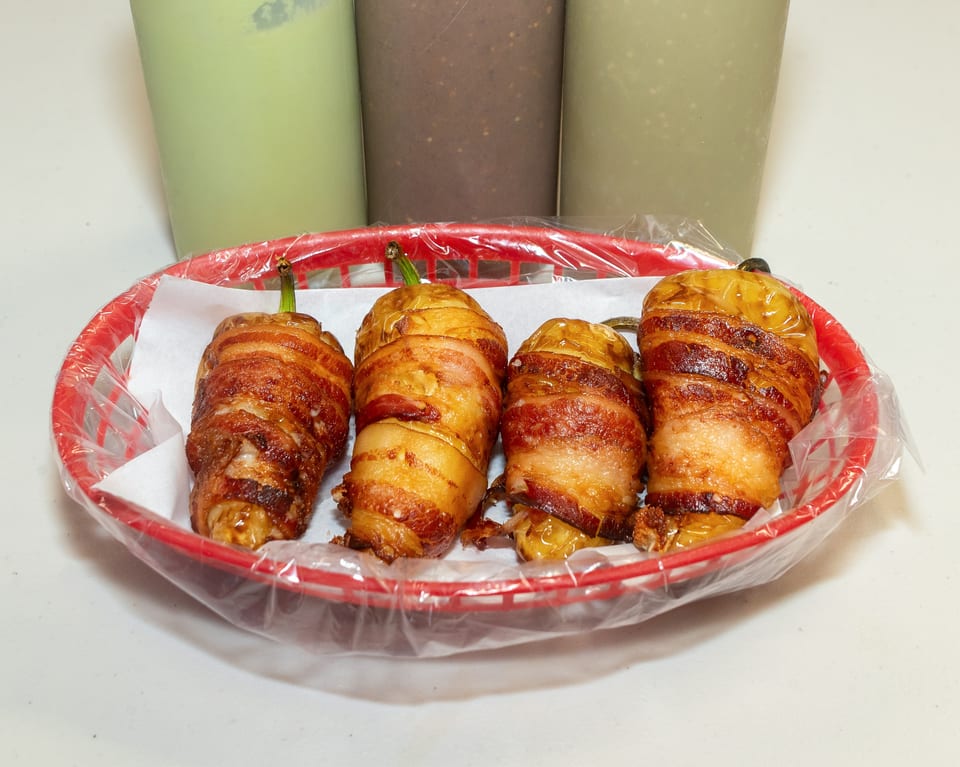 Four bacon-wrapped peppers in a red plastic basket with three drinks in the background.