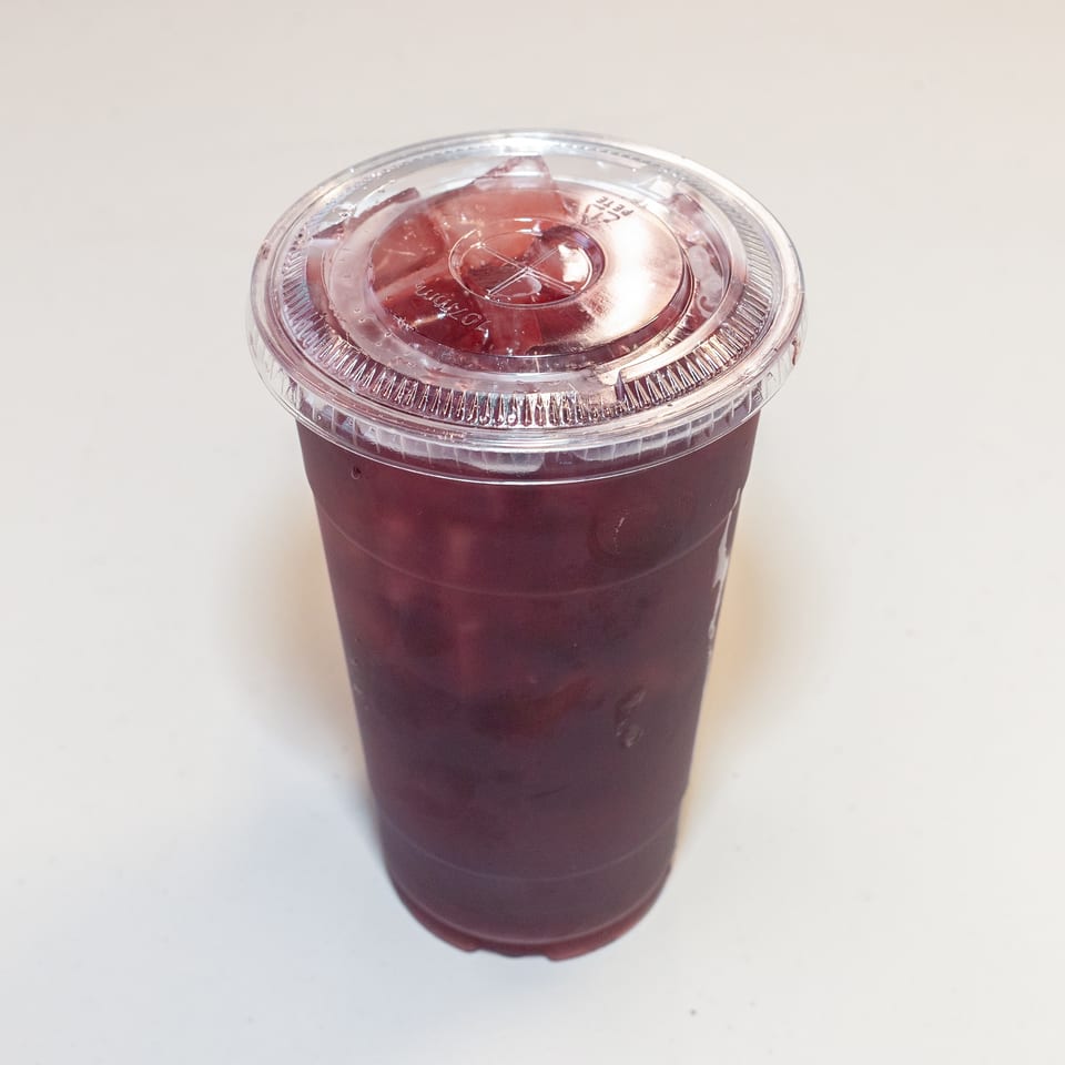 A clear plastic cup filled with a dark red beverage and ice, topped with a clear lid.