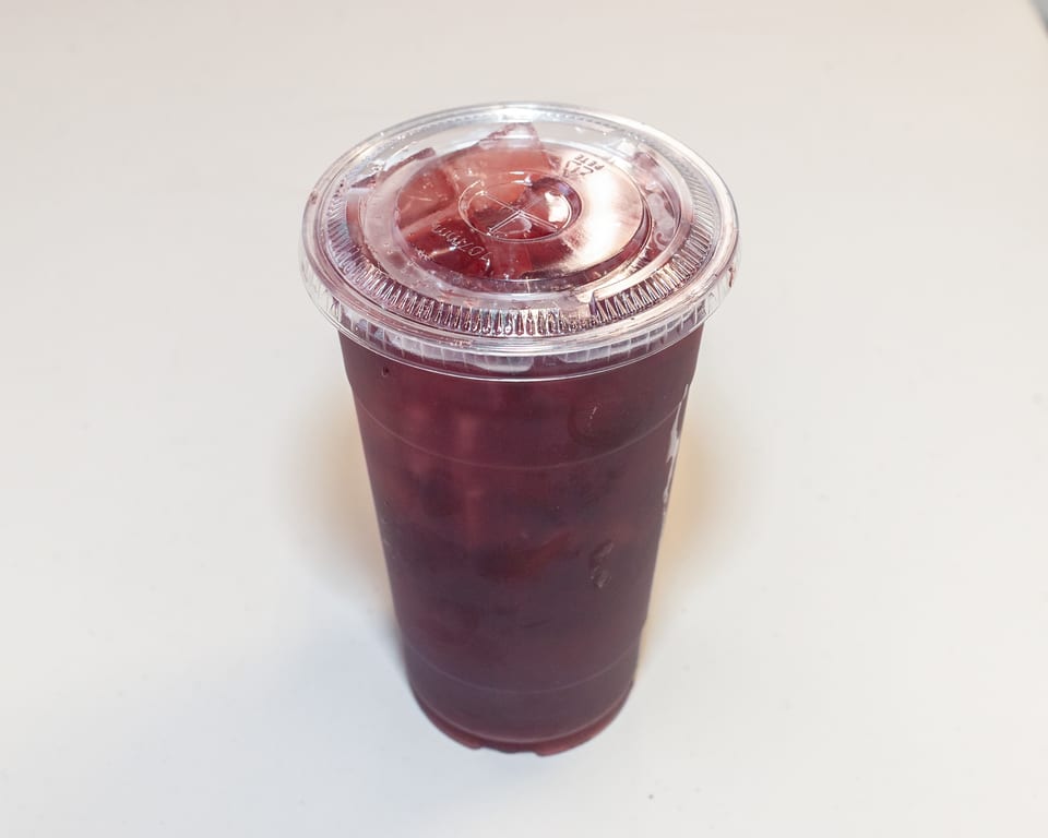A clear plastic cup filled with a dark red beverage and ice, topped with a clear lid.