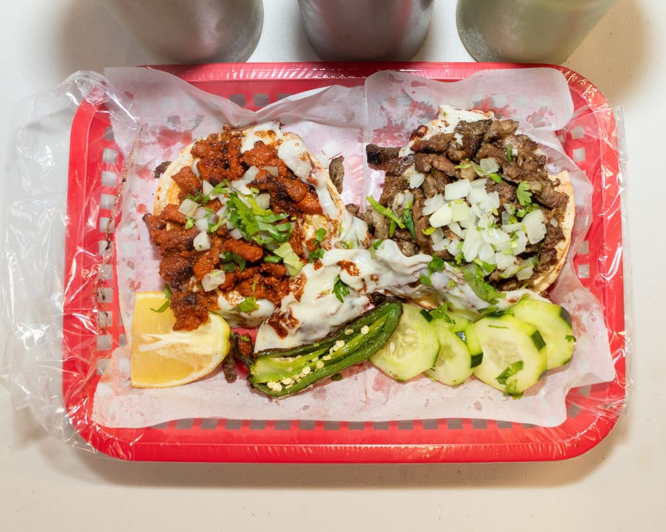 Two tacos, one al pastor and one carne asada, with grilled jalapeño, lemon, and cucumber slices.